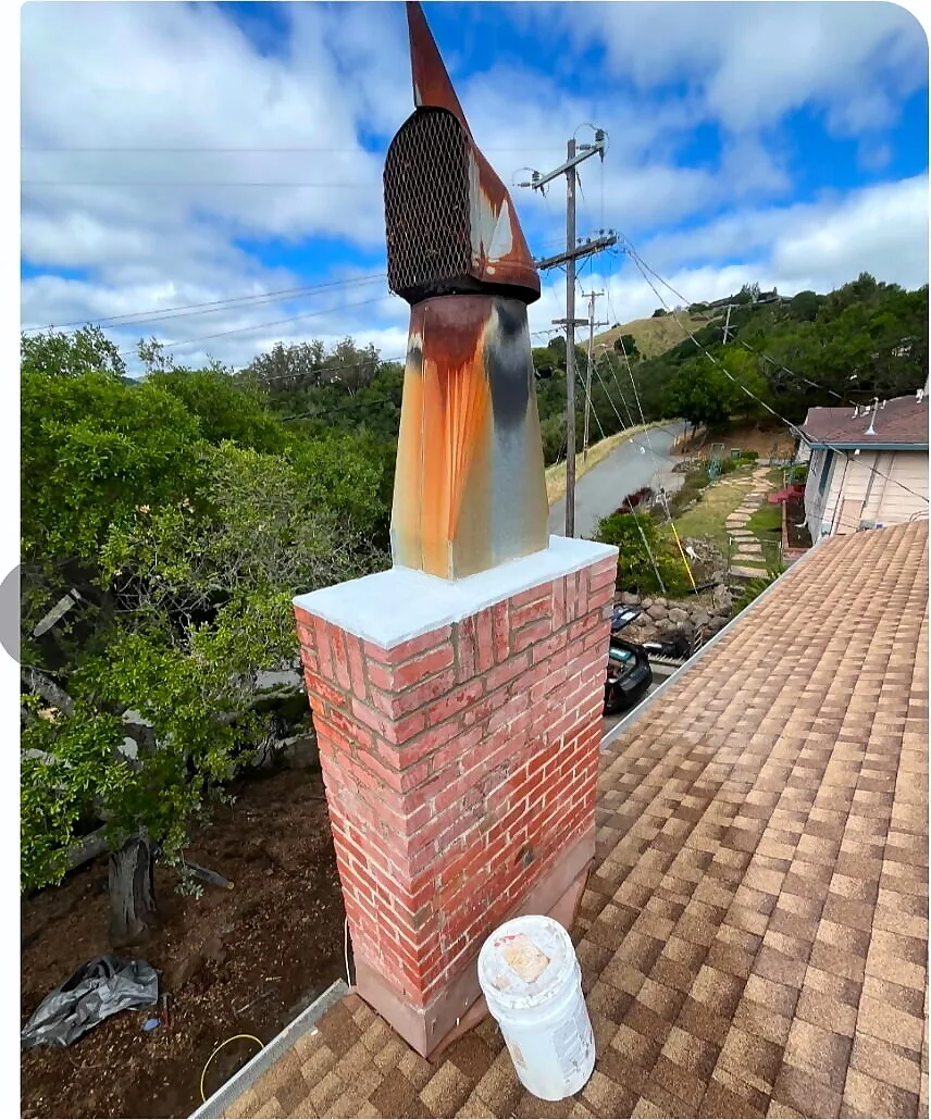 Brick chimney with a rusted spark arrestor cap on a shingle roof before replacement