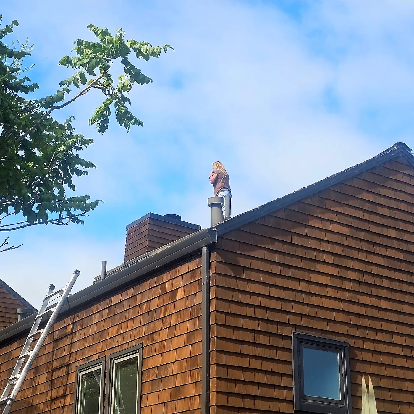 Heart's Chimney technician on a cedar shake roof beside a chimney with a ladder set below