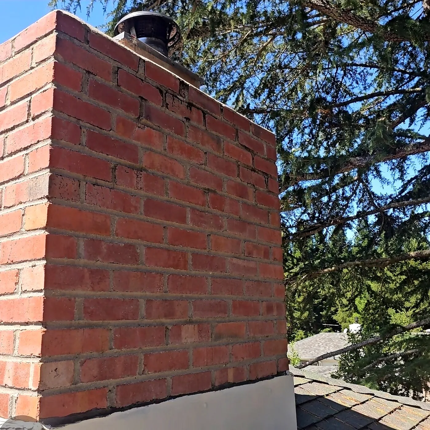 Clean red brick chimney with a round spark arrestor cap against a blue sky and cedar tree