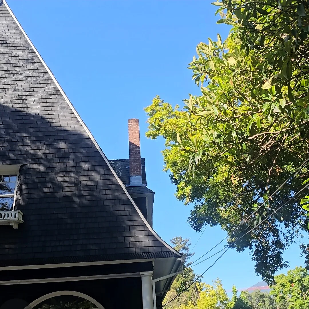 Steep shingled turret roof of a classic home with a brick chimney behind trees